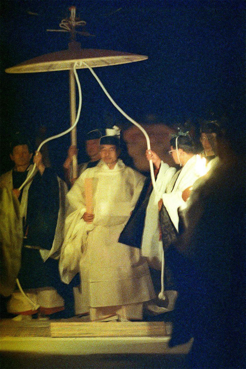 Акихито идет к помещению Сукидэн в сопровождении служащих Управления императорского двора, которые держат над ним зонт, украшенный изображением феникса. Снимок сделан 23 ноября 1990 года в восточной части императорского дворца. (Jiji Press)