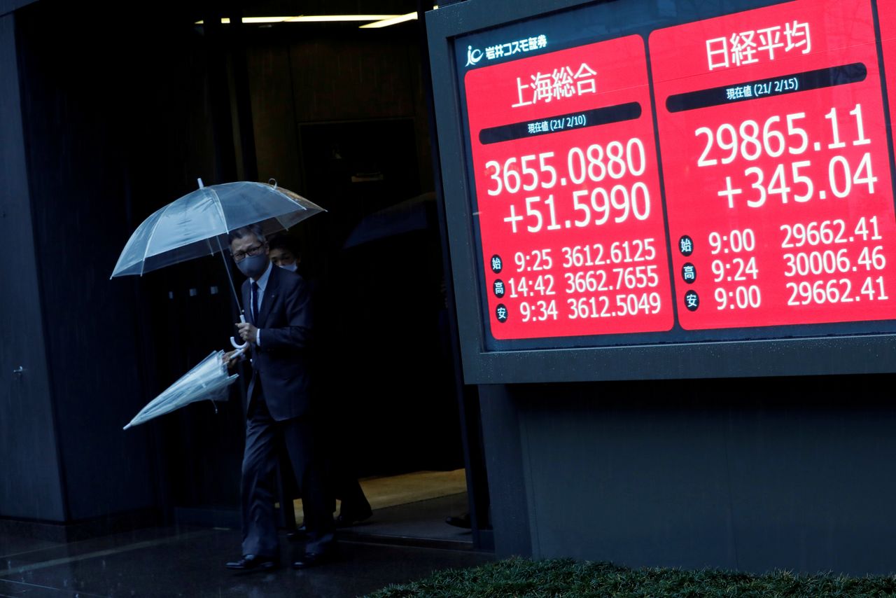 Men holding umbrellas walk near an electric board showing Nikkei index a brokerage in Tokyo, Japan February 15, 2021. REUTERS/Kim Kyung-Hoon