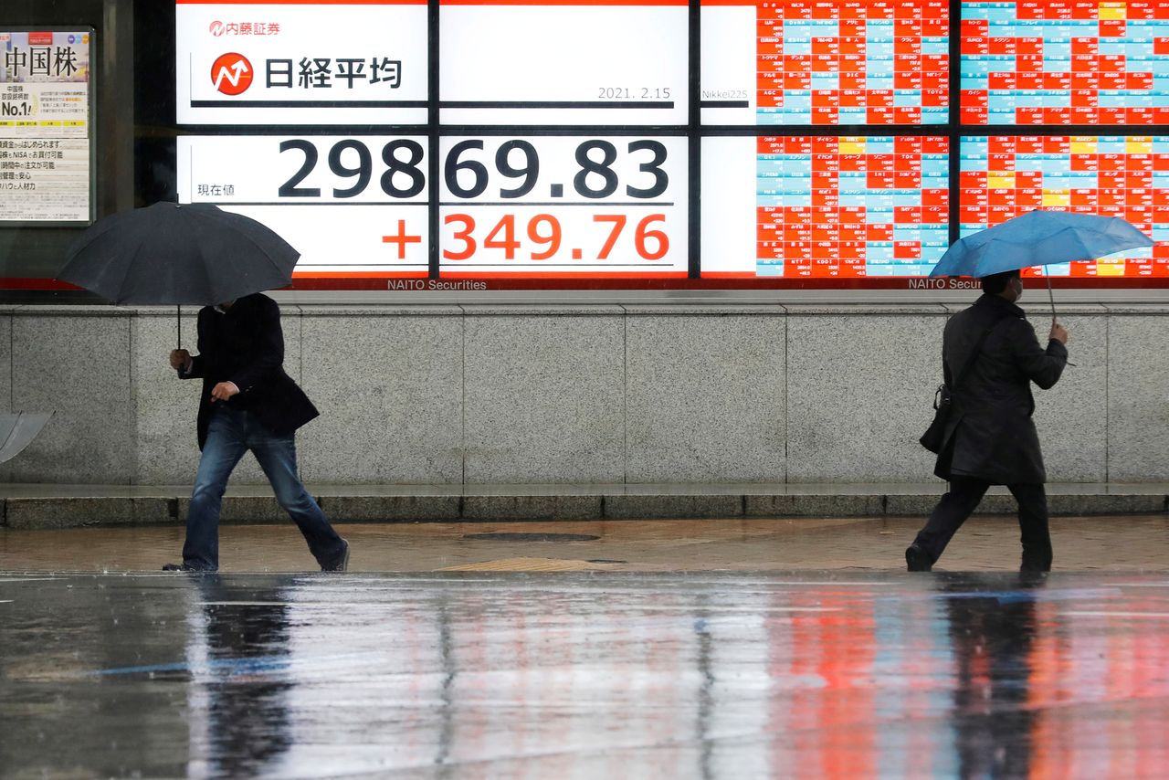 Men holding umbrellas walk in front of an electric board showing Nikkei index at a brokerage in Tokyo, Japan February 15, 2021. REUTERS/Kim Kyung-Hoon