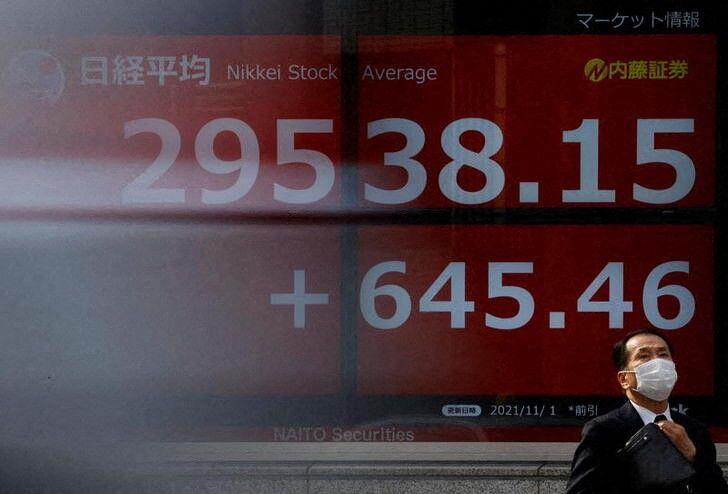 A man wearing a protective face mask stands in front of an electronic board showing Japan’s Nikkei share average, amid the coronavirus disease (COVID-19) pandemic, in Tokyo, Japan November 1, 2021. Picture taken with a slow shutter speed.  REUTERS/Issei Kato