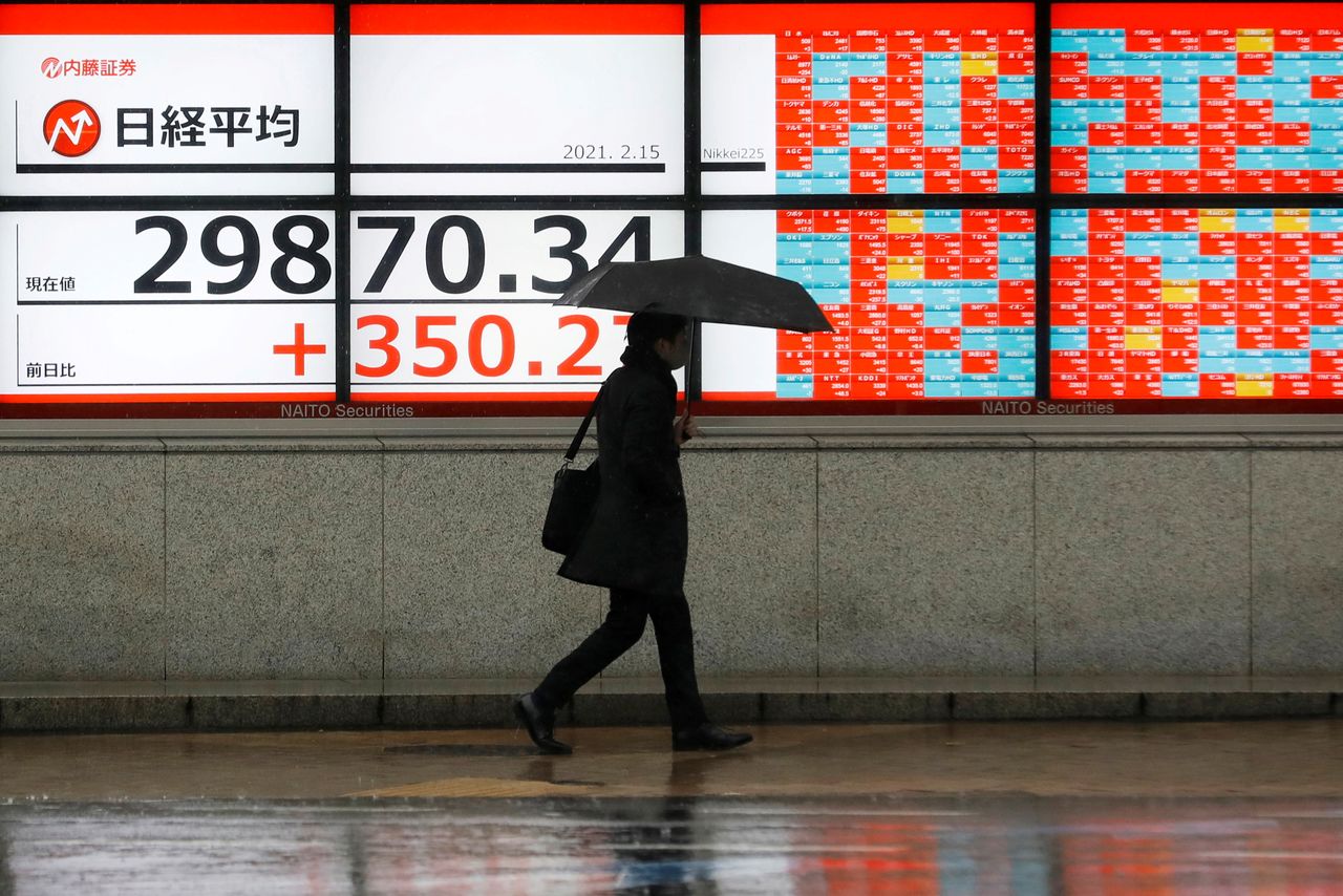 A man holding an umbrella walks in front of an electric board showing Nikkei index a brokerage in Tokyo, Japan February 15, 2021. REUTERS/Kim Kyung-Hoon